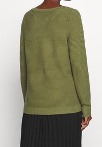 Sweter oliwkowozielony o teksturowanym wzorze dzianiny, z okrągłym dekoltem, długimi rękawami i ściągaczami na mankietach. Noszony na plisowanej czarnej spódnicy.