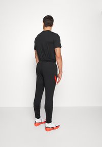 Zwart sporttop met korte mouwen, gecombineerd met zwarte joggers met een rode accent. G gedragen met witte en oranje sportschoenen.