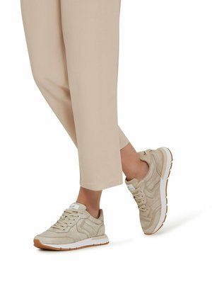 Beige suede sneakers with a white sole, textured side accents, and lace-up design. Worn with cropped beige pants, displaying a casual look.