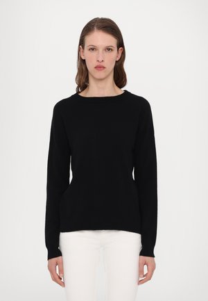 Schwarzer Strickpullover mit langen Ärmeln und Rundhalsausschnitt, der eine glatte Textur und eine lockere Passform aufweist. Über weißen Hosen getragen.