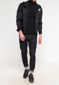 Svart lättviktig jacka med huva och logotyp, lagd över en svart polotröja. Mörka jeans och kamouflagemönstrade sneakers, med uppvikt ärm och strukturerad tyg.