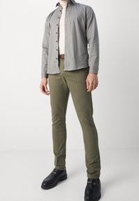 Chemise à motifs gris avec boutons, sous-vêtement crème, pantalon slim vert olive, et mocassins noirs avec détails dorés.