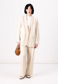Beige kavaj med två framfickor, ihop med vida beige byxor. Modellen håller en brun vävd handväska och bär svarta och vita skor.