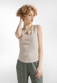 Joven con cabello rizado, vestida con una blusa sin mangas beige y pantalones grises, de pie con una mano en el cuello, mirando hacia abajo.