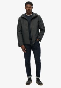 Donkergrijze jas met een ritssluiting aan de voorkant, twee borstzakken en een capuchon. Draagt over een marineblauw sweatshirt en donkere spijkerbroek. Zwarte schoenen met veters.