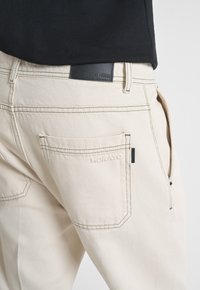 Pantalones de algodón beige con costuras en verde en contraste. Presenta una etiqueta de cuero negro con "ANTONY MORATO" en la cinturilla y un bolsillo lateral.