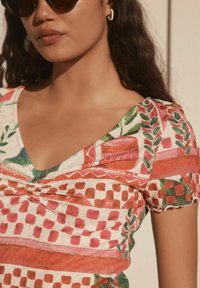 Kurzärmliges Oberteil mit V-Ausschnitt, das mit einem Muster aus roten, grünen und pinkfarbenen Formen, strukturiertem Stoff und einem gerafften Detail am Ausschnitt versehen ist.