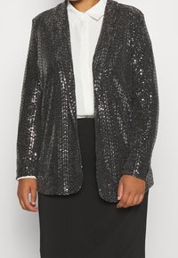 Persona che indossa un blazer nero di paillettes sopra una camicia bianca con colletto e una gonna nera, in piedi contro uno sfondo semplice.