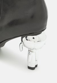 Chaussure noire avec un talon transparent en forme de petit robot, présentant des jambes, des bras définis et un détail de visage semblable à une visière, sur fond blanc.
