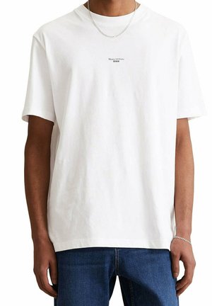 Homme portant un t-shirt blanc Marc O’Polo Denim et un jean bleu, accessoirisé avec un collier chaîne en argent et un bracelet.