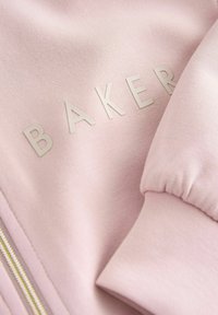 Roze stof met verhoogde beige letters die "BAKER" spellen en een gerimpelde manchette nabij een gouden rits.