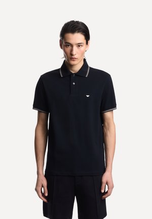 Giovane con capelli neri e lisci indossa una polo nera a maniche corte con bordi bianchi e un piccolo logo sul petto.