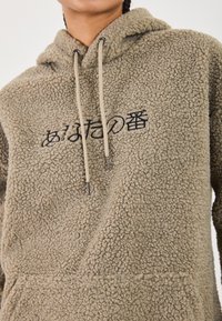 Hoodie en polaire beige avec une surface texturée, comportant une poche frontale, des cordons de serrage ajustables et un design brodé noir.