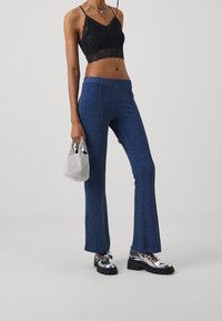 Leggings évasés bleus pailletés avec une texture lisse, associés à un crop top en dentelle noire et des chaussures à semelles compensées argentées. Tenant un petit sac en maille.