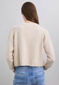 Mujer con cabello largo y castaño que lleva un suéter de punto crema y jeans azul claro, de espaldas sobre un fondo gris liso.