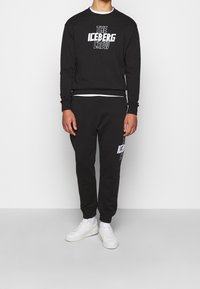 Felpa nera con grafica "THE ICEBERG CREW" sul davanti, abbinata a joggers neri con dettaglio del logo e sneaker bianche.