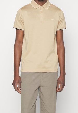 Polo beige en coton, avec une patte à deux boutons, un bord contrastant noir sur les manches, et un logo discret sur la poitrine.