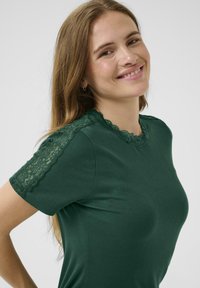 T-shirt vert côtelé avec dentelle sur les épaules et le col. Conception ajustée et texture douce, avec un col rond et des manches courtes.
