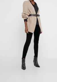Gilet en tricot beige avec des manches retroussées, ceinturé à la taille, associé à des leggings en velours noir et des bottines noires à talons pointus.