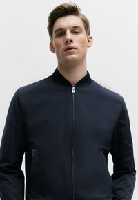 Navy bomber jacket with a high collar, front zip closure, and side pockets. Smooth, lightweight fabric with a minimalist design.