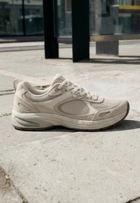 Baskets de sport beiges avec panneaux en maille et en daim reposant sur un trottoir en béton à l'extérieur, près d'un abri de bus en verre.