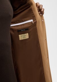 Interieur van een bruine wollen jas met een gladde bruine voering, een ritszak en labels met de tekst "GREENWOOL" en "FINEST WOOL."