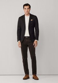 Hombre vestido con blazer negro, camisa blanca, pantalones oscuros, cinturón marrón y zapatos de vestir marrones de pie contra un fondo liso.