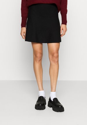 Person iført en sort mininederdel, hvide sokker og kraftige sorte plateau-sko, stående mod en ensartet lys baggrund.