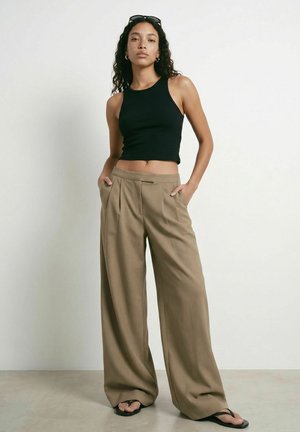 Femme aux longs cheveux bouclés porte un crop top noir sans manches, un pantalon large beige, des tongs noires et des lunettes de soleil sur la tête, debout les mains dans les poches.