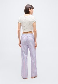 Femme debout pieds nus, vue de dos, portant un haut court blanc à motifs et un pantalon ample lavande à rayures bleues verticales.