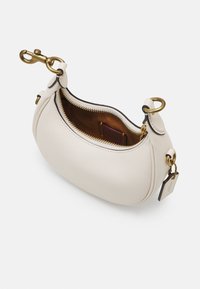 Petit sac à main en cuir blanc incurvé avec fermeture éclair et fermoirs dorés, partiellement ouvert montrant l'intérieur marron et une étiquette en cuir suspendue.