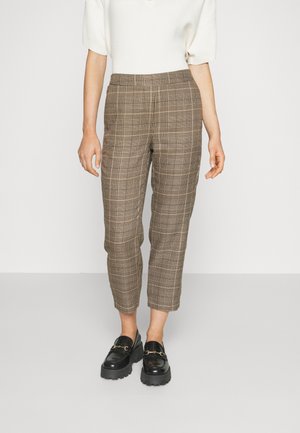 Personne portant un pantalon court marron à carreaux, un haut blanc à manches courtes et des mocassins noirs épais, debout sur un sol gris.