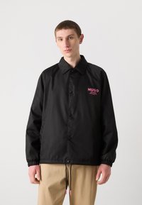 Črna vetrovka z gumbi spredaj, ki ima roza "HUGO" logo na prsih in elastične manšete. Vkljčuje tudi zategovalno rob.L