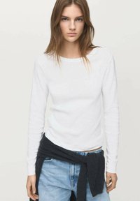 Cămașă albă cu mâneci lungi din bumbac, asociată cu blugi albaștri. Un pulover gri închis este legat în jurul taliei. Design simplu, ajustat.