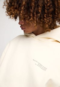 Felpa crema con grande cappuccio, tessuto morbido, marchio ricamato sul davanti, vestibilità rilassata e spalle scese per una maggiore comodità.