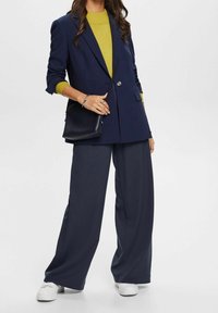 Blazer surdimensionné bleu marine sur un pull moutarde, associé à un pantalon large bleu marine et des baskets blanches. Un petit sac noir complète la tenue.