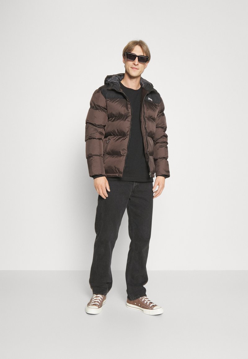 Veste matelassée marron avec capuche, présentant un empiècement noir, des poches zippées et un design matelassé, portée avec un pantalon noir et des baskets marron.