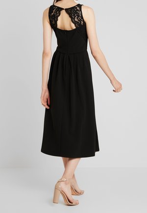 Frau trägt ein ärmelloses schwarzes Midikleid mit Spitzendetail am oberen Rücken und hautfarbene High-Heel-Sandalen, steht vor einem grauen Hintergrund.