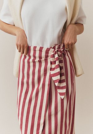 Frau trägt weißes Hemd mit cremefarbenem Weste, die an den Schultern gebunden ist, und rot-weiß gestreiften Wickelrock mit Knoten vorne.