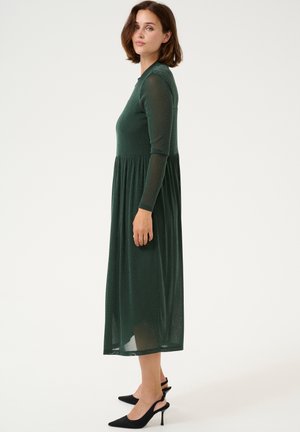 Sateen vihreä pitkähihais dressi, jossa on tiukka pääntie ja virtaava hame, valmistettu läpikuultavasta materiaalista, yhdistettynä mustiin teräväkärkisiin korkokenkiin.