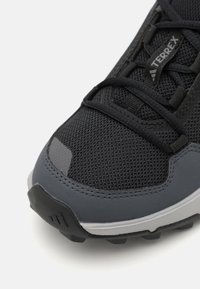 Black athletic shoe with mesh upper, gray accents, textured rubber sole, and a reinforced toe cap. Features lacing and a logo on the side.