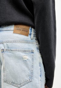 Svijetloplave traperice s istrošenim detaljima, koje imaju smeđi kožni etiketu s natpisom "PULL&BEAR DENIM GOODS" na struku.