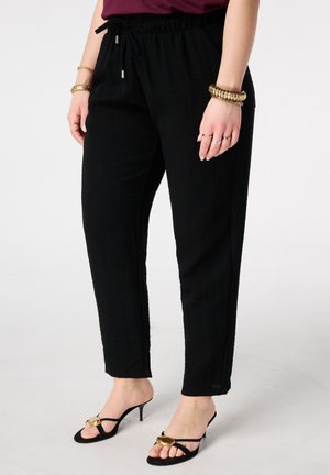 Vrouw draagt zwarte broek met trekkoord en zwarte sandalen met hoge hak, versierd met gouden ovale details, en gouden armbanden en ringen aan de handen.
