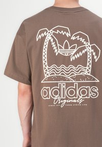 Hnědé bavlněné tričko s bílou grafikou palem a vln, se textem "adidas Originals" v tučném písmu ve spodní části.