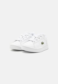 Vita sneakers i slät syntetiskt material med rund tå, snörningsdesign och liten grön logotyp på utsidan. Gummisula.