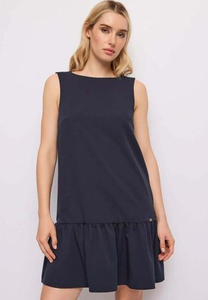 Donna bionda che indossa un vestito senza maniche color blu navy scuro con orlo arricciato, in piedi davanti a uno sfondo chiaro uniforme.