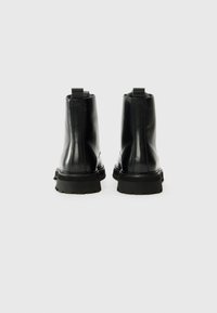 Par de botas negras de cuero hasta el tobillo con suelas de goma gruesa, vistas desde la parte de atrás sobre un fondo claro liso.