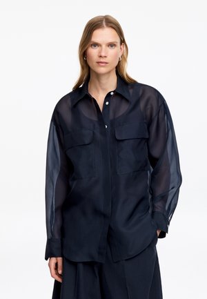 Femme aux cheveux blonds portant un chemisier noir transparent à manches longues avec des poches sur le devant, debout devant un fond blanc.