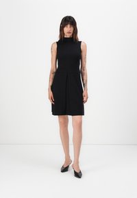 Black sleeveless dress with a high neck, pleated skirt, and smooth fabric. Paired with black block-heel shoes.
