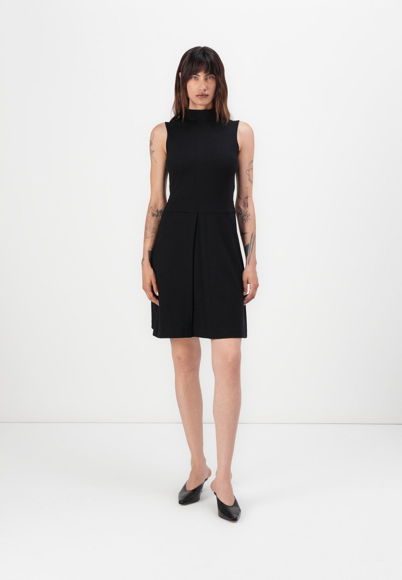Black sleeveless dress with a high neck, pleated skirt, and smooth fabric. Paired with black block-heel shoes.